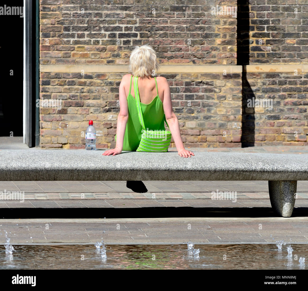 Young woman in a lime green dress relaxing by water in Granary Square, Parkside, King's Cross, London, England, UK. Stock Photo