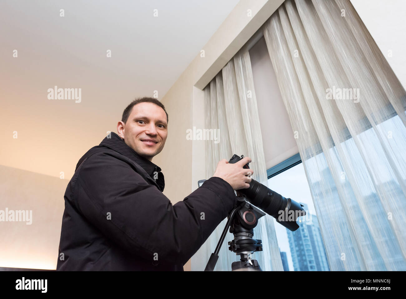 Young happy smiling man photographer with tripod taking pictures inside room of hotel, apartment building through window in New York City, NYC urban Stock Photo