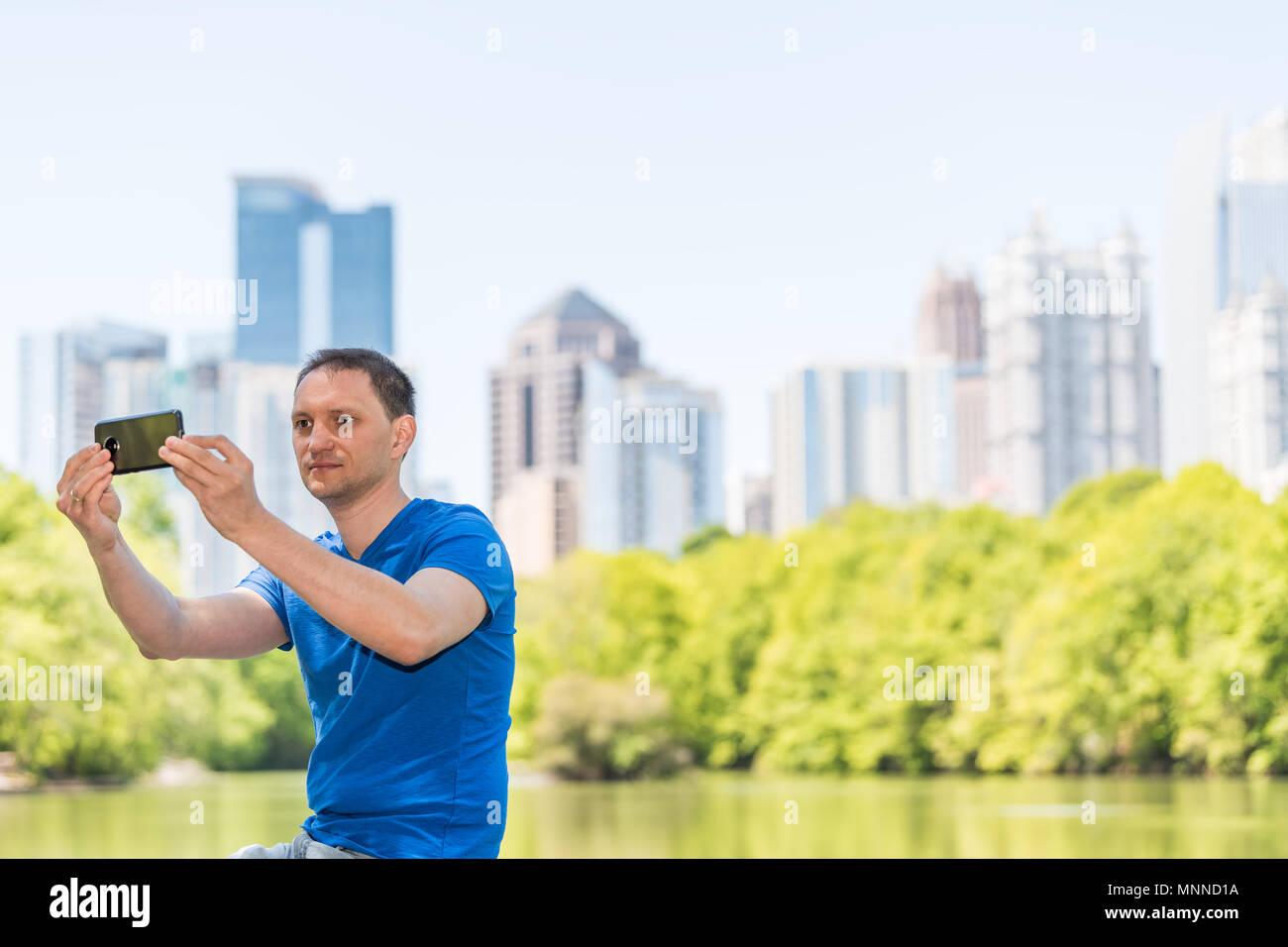 Young man sitting taking selfie in Piedmont Park in Atlanta, Georgia with scenic water, and cityscape skyline of urban city skyscrapers downtown, Lake Stock Photo