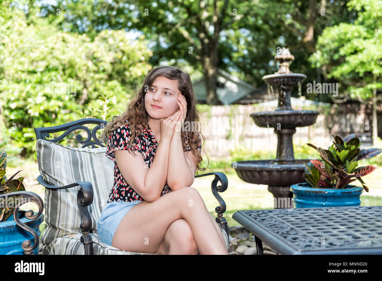 Young woman sitting on patio chair in outdoor spring garden in backyard porch of home happy smiling zen with fountain, table, plants Stock Photo