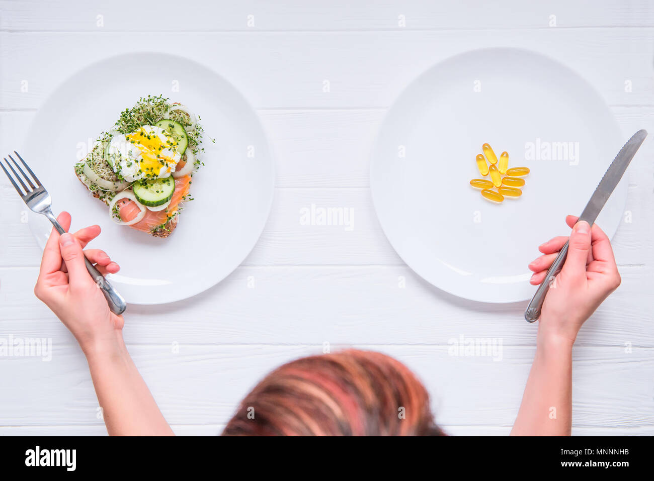 Top view woman with cutlery in hands choosing between healthy food or medical pills on the white plates and wooden table. Choice between natural and synthetic way of health care. Alternative medicine. Stock Photo