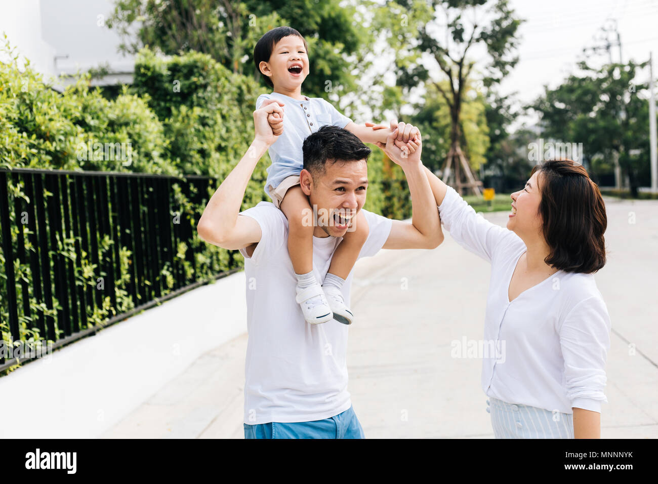 Cute Asian father piggybacking his son along with his wife in the park. Excited family raising hands together with happiness Stock Photo