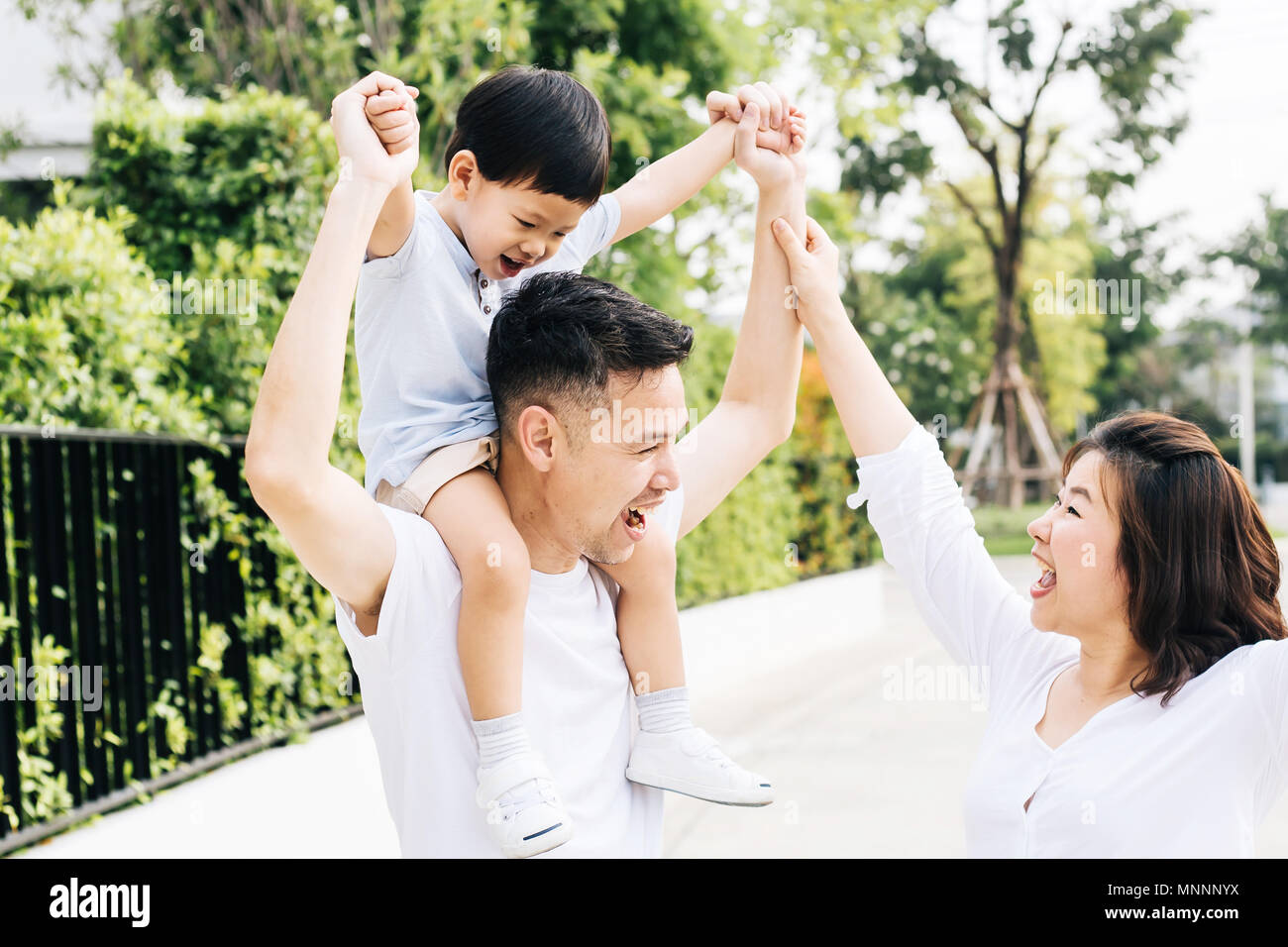 Cute Asian father piggybacking his son along with his wife in the park. Excited family raising hands together with happiness Stock Photo