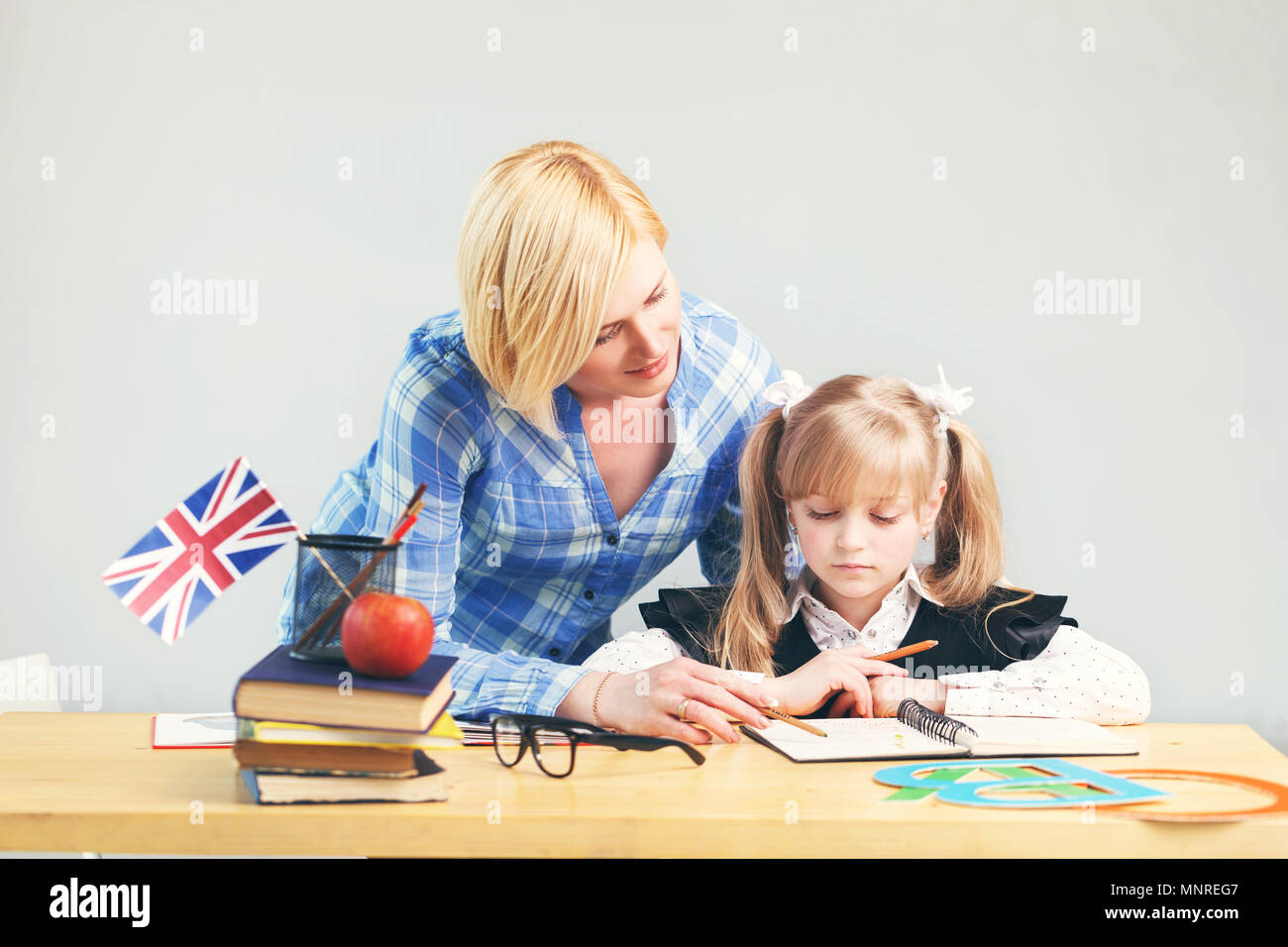 Pretty adult female teacher helps kid girl to learn English language, school table with books, flag and letters in light classroom Stock Photo