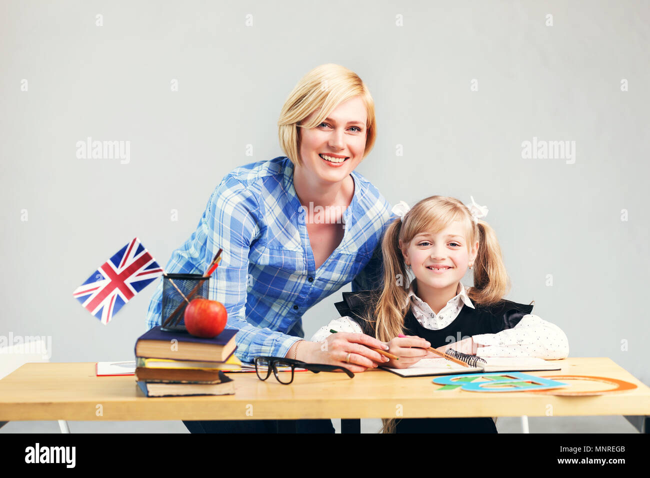 Woman educator helps kid girl to learn English language, school table with books, flag and letters in light classroom Stock Photo