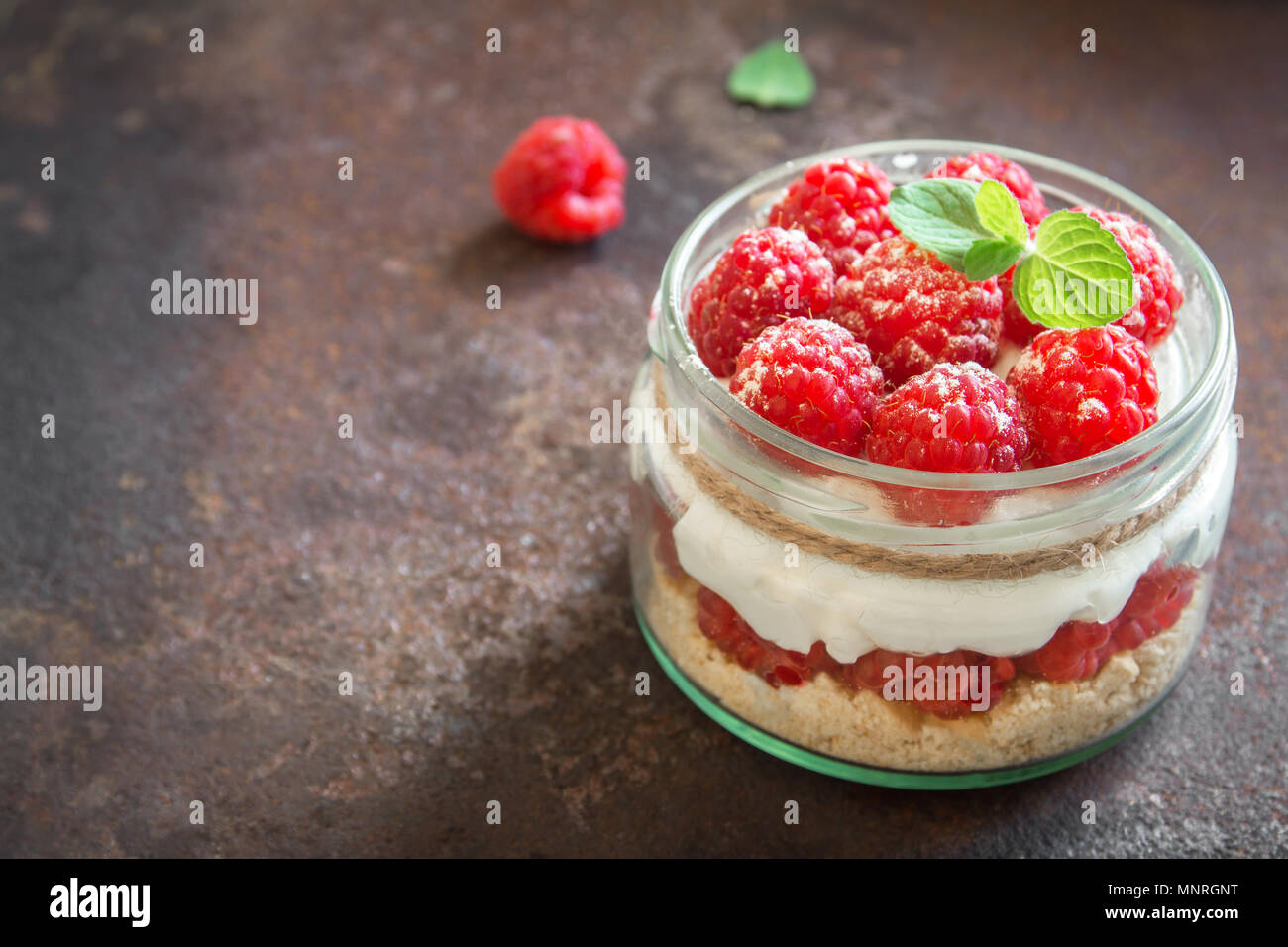 Raspberry cheesecake in glass jar with fresh raspberries and cream cheese  on rustic metal background. Healthy homemade summer berry layered dessert. Stock Photo