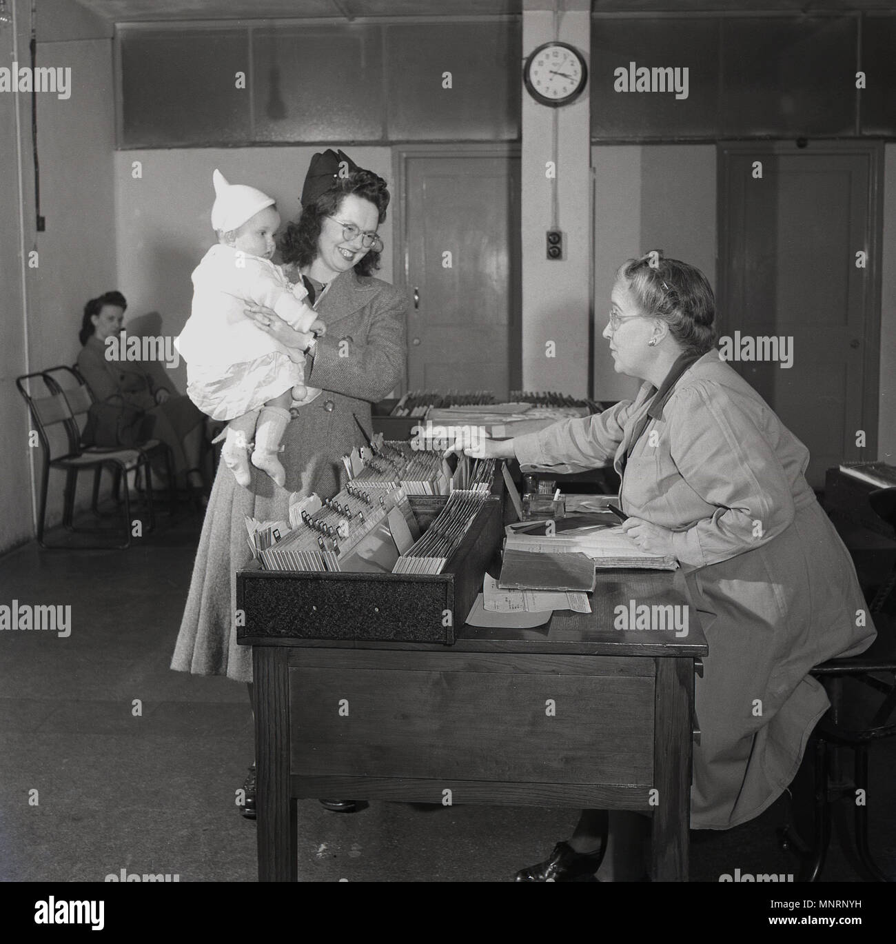 1950s, historical, happy lady with her infant child, talking to the elderly female receptionist sitting at desk at a Doctor's surgery, London, England, UK. Stock Photo