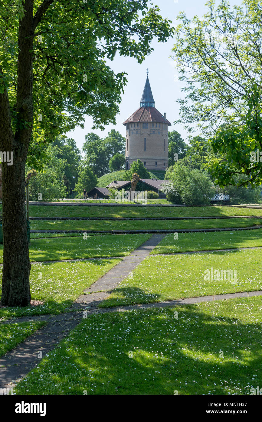 Narrow tower with red tile roof and green spire in lush park of neatly mown grass and concrete path. Stock Photo