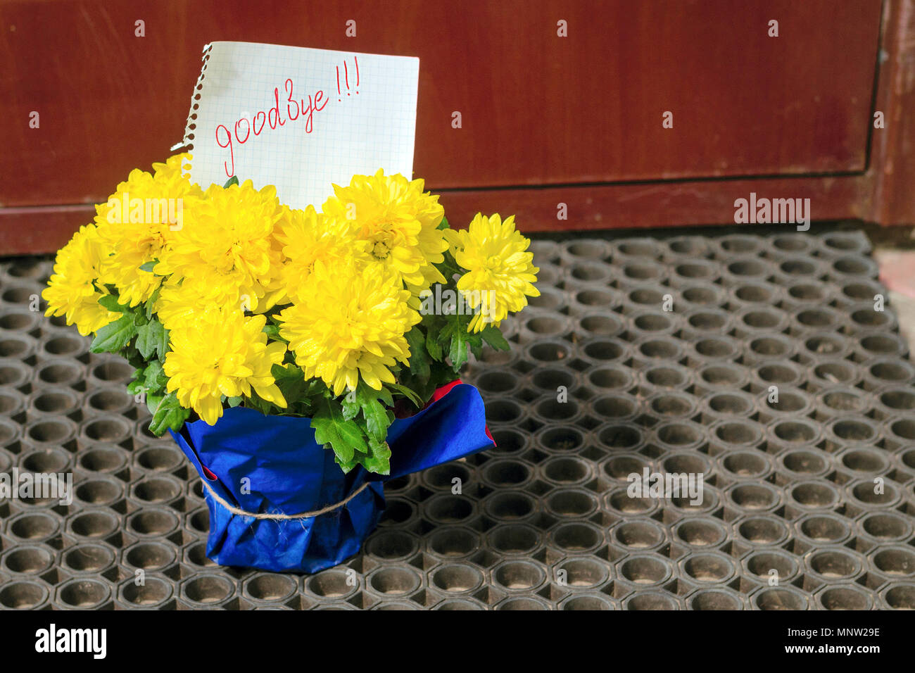 A letter on a piece of paper 'Goodbye' is in a bouquet of yellow flowers at the door of the house. The concept of parting. Close-up. Stock Photo