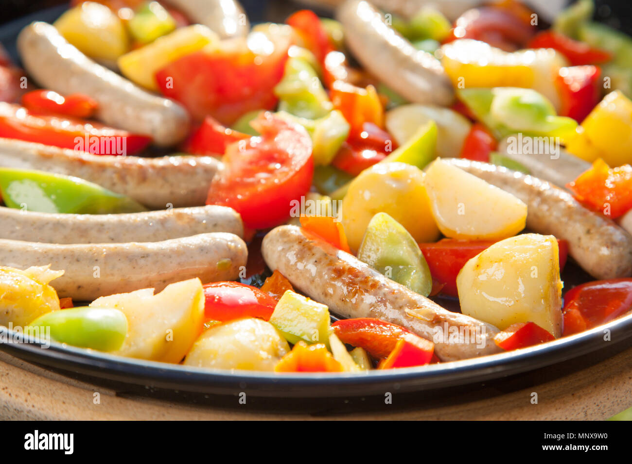 small sausages, potatoes and tomatoes in a hot pan coooking outside in the sun Stock Photo