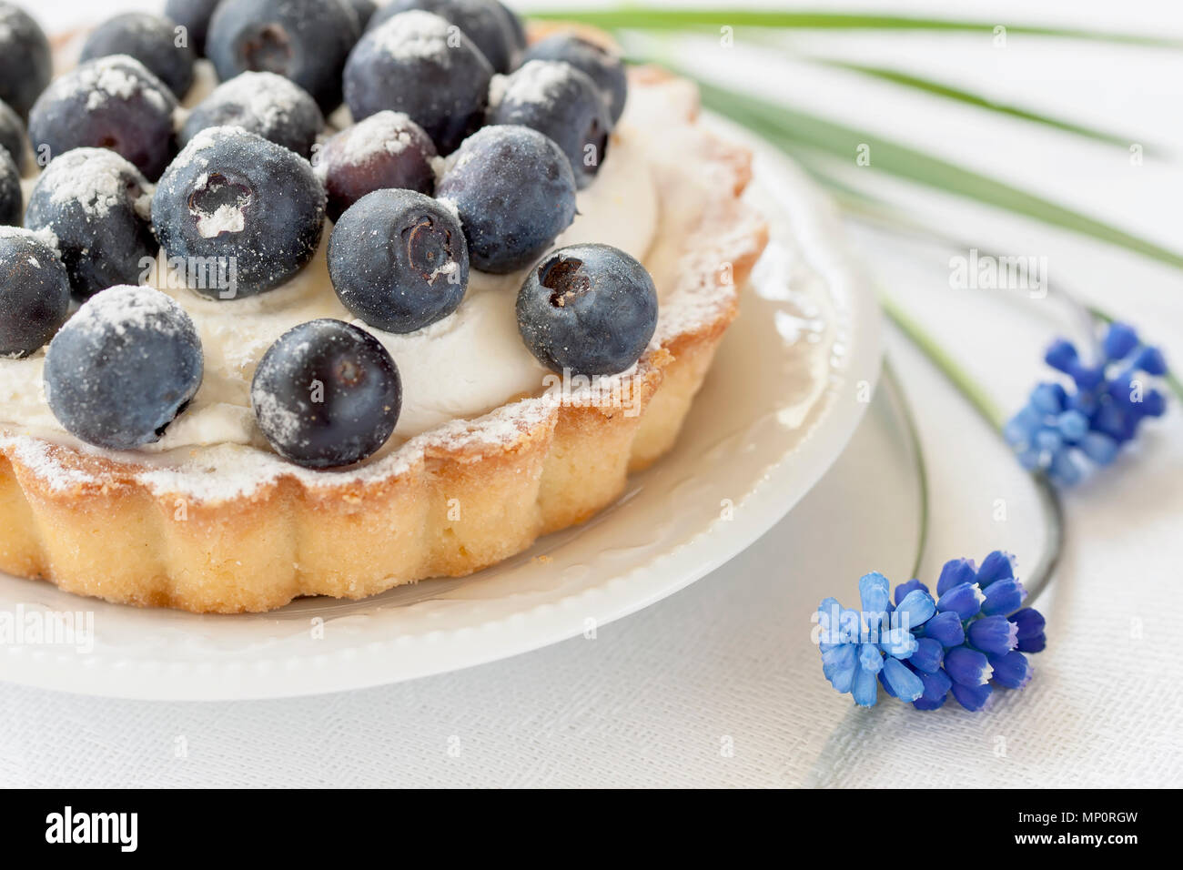 Fresh blueberry tart with vanilla custard in portion plate close-up, next to elegant blue flowers. Delicious and romantic summer berry dessert. Stock Photo