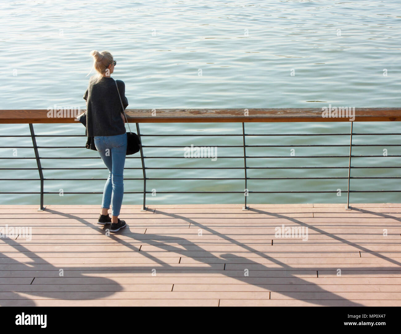 Belgrade, Serbia - October 15, 2017 : Young blonde woman standing  on the river promenade leaning on the railings and looking at water, from behind Stock Photo