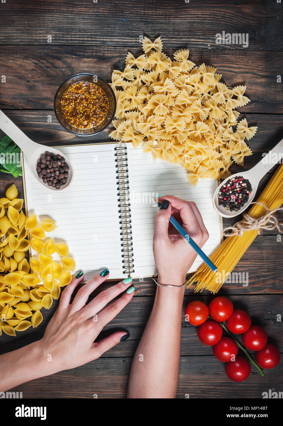 Female hands writing notepad. spices, pasta and vegetables around notebook on wooden table. Mockup. Stock Photo