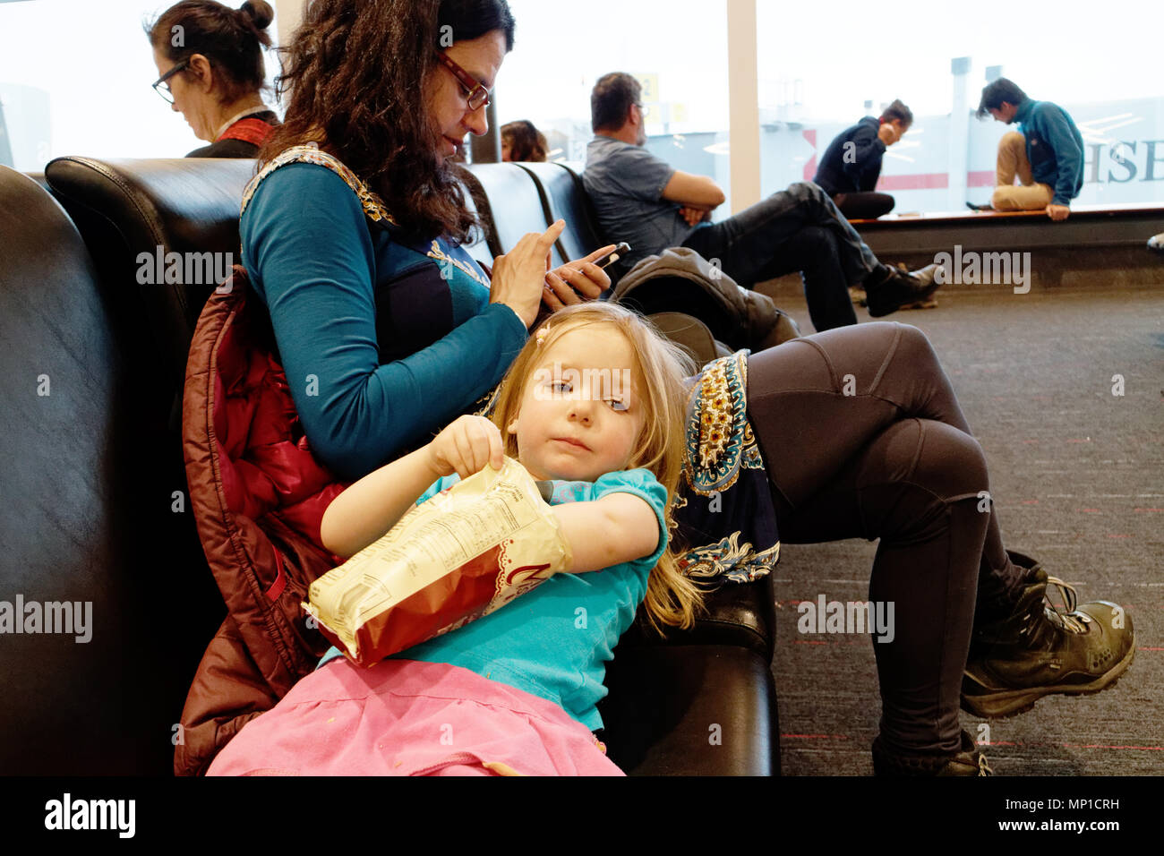 A mother using her iphone while her daughter relaxes, leaning back on her thigh eating a bag of crisps while waiting for their flight to be called Stock Photo