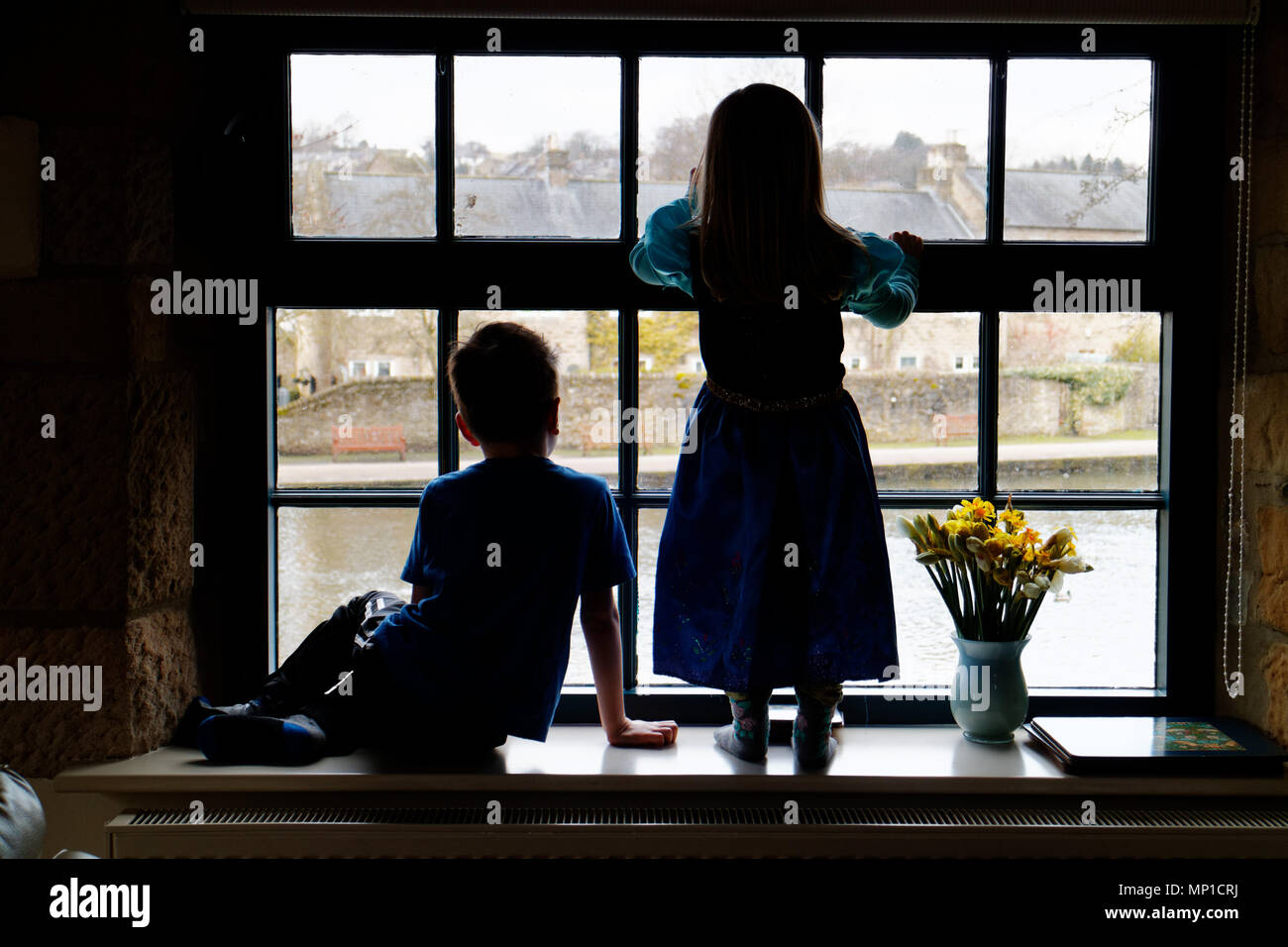 Brother (5 yrs old) and sister (3 yrs old) sat on a window sill, silhouetted against a sash window with the River Wye in Bakewell outside Stock Photo