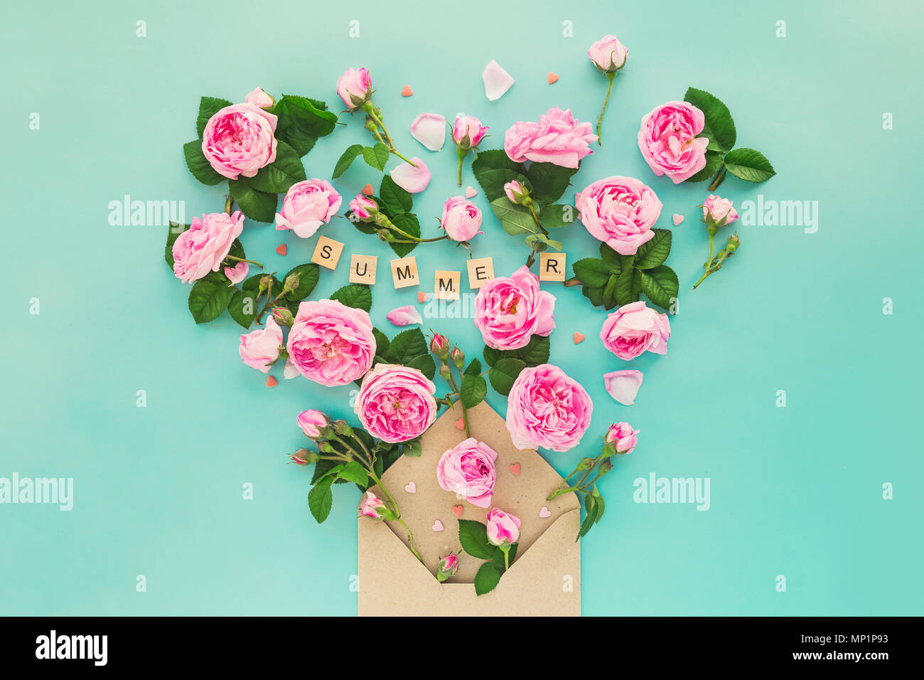 Creative layout with pink tea rose flowers, leaves fly out of croped craft paper envelope in shape of heart on the light turquoise background. Word Summer lettering by wooden letters. Space for text. Stock Photo