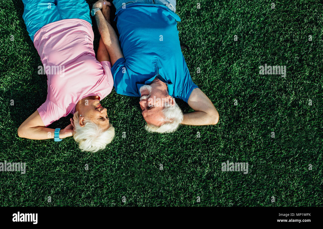Senior couple laying in grass, top view. Elderly man and woman lying on grass together and hold their hands Stock Photo