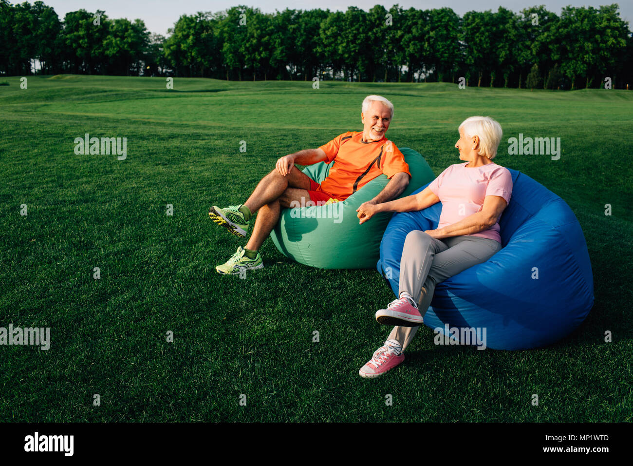 Senior people, elderly man and woman sitting on beanbags on grass, looking at each other and laughing, Stock Photo