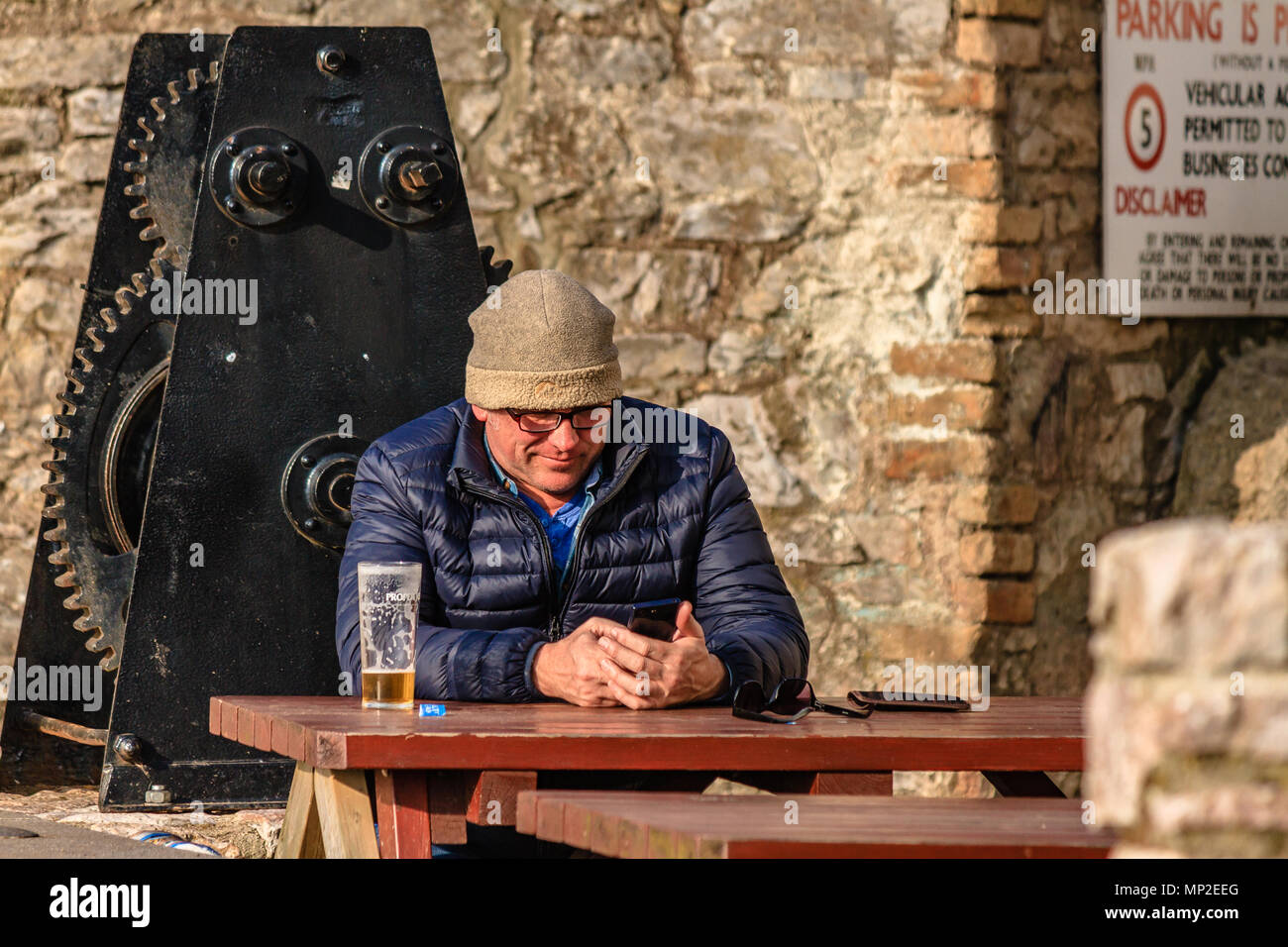 Man in warm coat and hat sat outside a pub with a nearly finished pint of beer and using a mobile phone. Teignmouth, Devon. Feb 2018. Stock Photo
