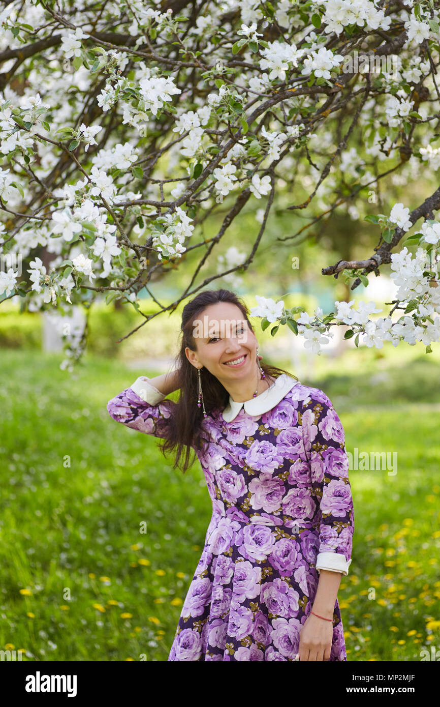 Beautiful brunette female wearing pretty dress with floral print standing near blooming apple tree Stock Photo