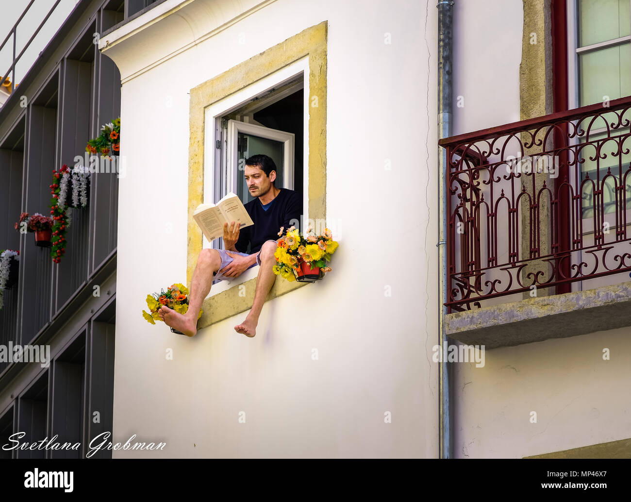 Man sitting on the window seal of an open window reading; man's legs dangling out of window Stock Photo