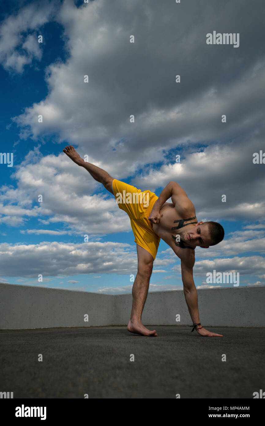 Tricking on street. Martial arts. Man makes kick with support on arm and leg barefoot. Shooted from bottom foreshortening against sky. Stock Photo