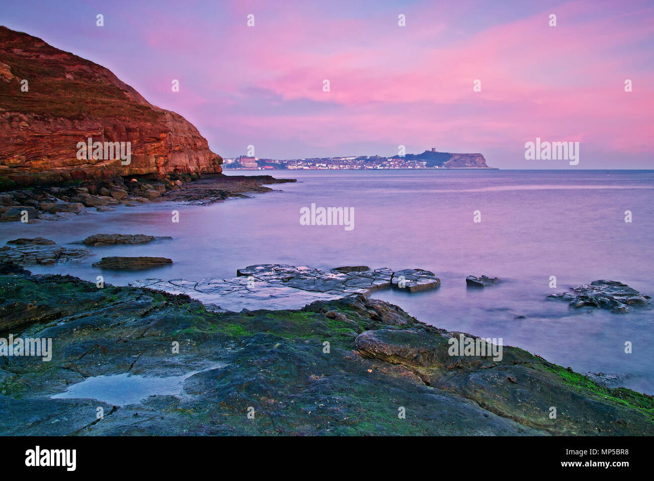 Dawn breaking over Scarborough Castle headland and an incoming tide, with views northwards over the North Sea from a rock pool in Cornelian Bay. Stock Photo