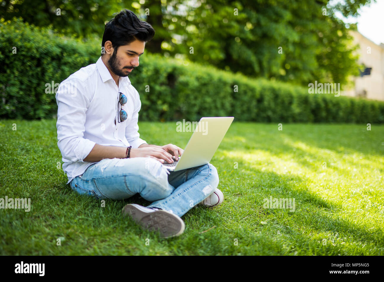 Young indian Student with a laptop sitting on the grass. Urban style. Stock Photo