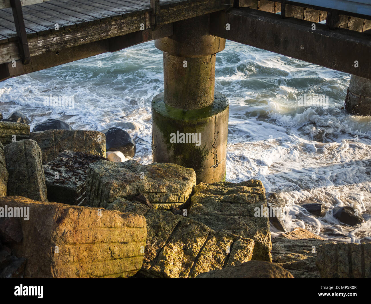 Concrete pillar of wooden jetty with rocks and waves during sunet Stock Photo