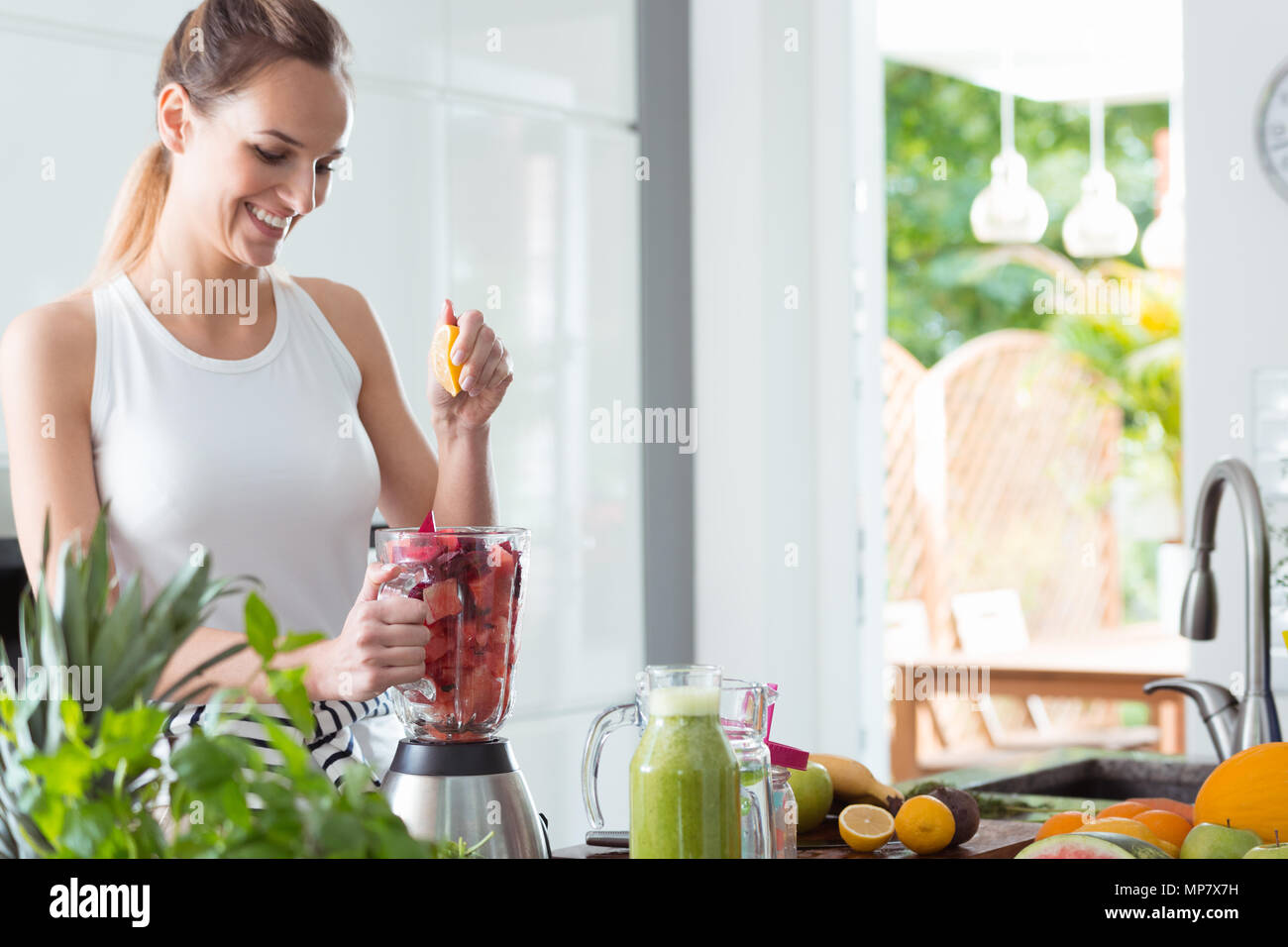 Smiling woman squeezing orange juice into blender with sliced watermelon and beet while preparing smoothie in kitchen Stock Photo