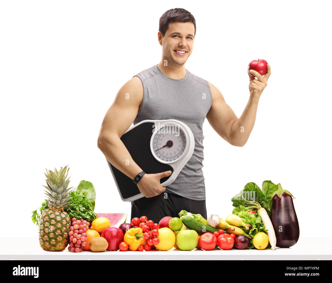 Young man in sportswear holding a weight scale and an apple behind a table with fruit and vegetables isolated on white background Stock Photo