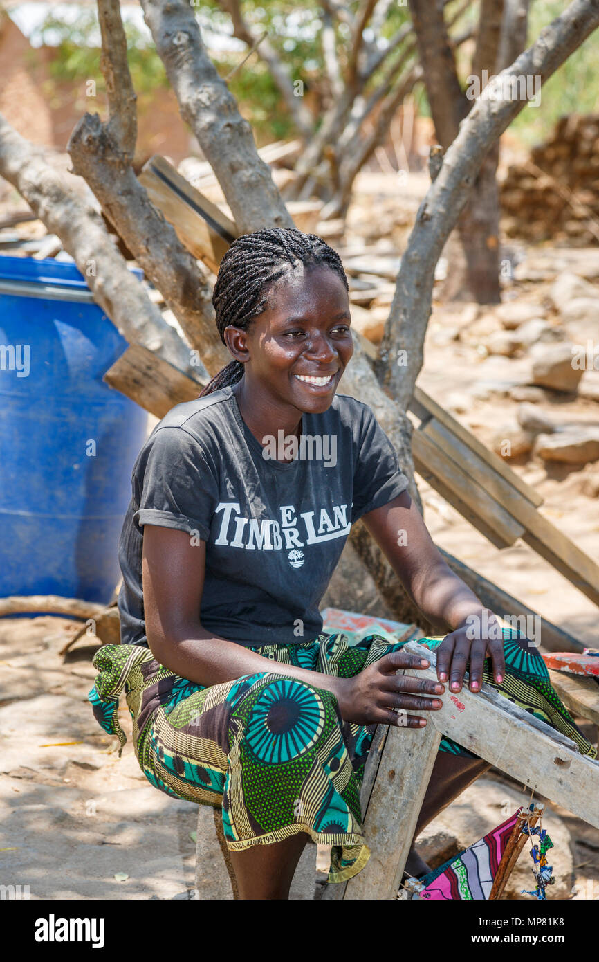 Smiling local African woman with braided hair sitting working outdoors at Katundu creative trade workshop, Likoma Island, Lake Malawi, Malawi, Africa Stock Photo