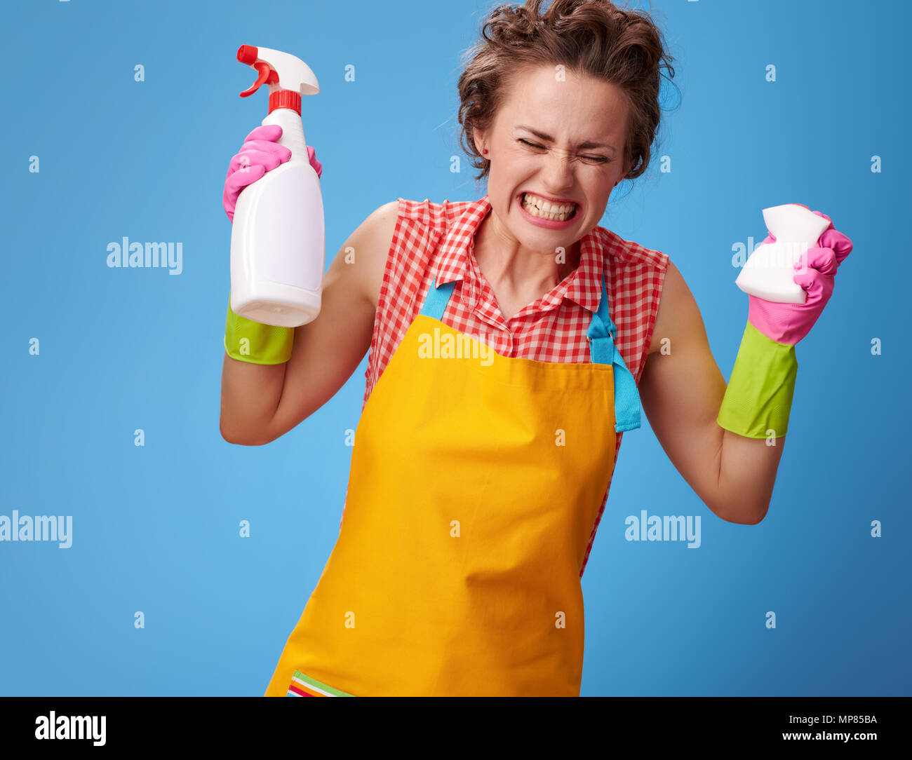 Big cleaning time. angry modern woman in a yellow apron with kitchen sponge and a bottle of detergent isolated on blue Stock Photo