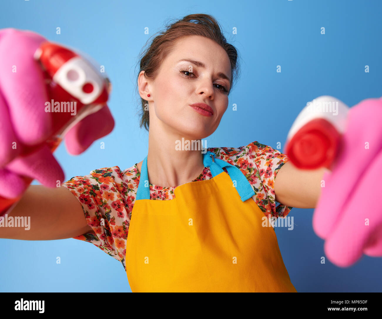 Big cleaning time. cool young housewife with rubber gloves using bottles of cleaning detergent as guns isolated on blue Stock Photo