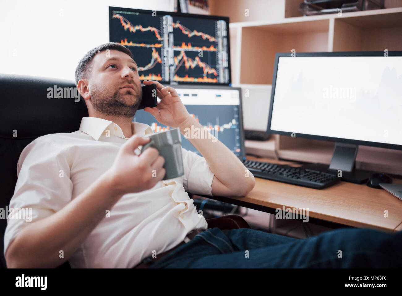 Over the shoulder view of and stock broker trading online while accepting orders by phone. Multiple computer screens ful of charts and data analyses in background Stock Photo