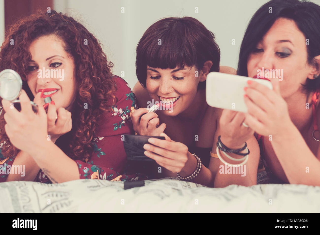 three females friends stay together lay down on the bed doing make up before go out to have party night. friendship concept one of them use a mobile p Stock Photo