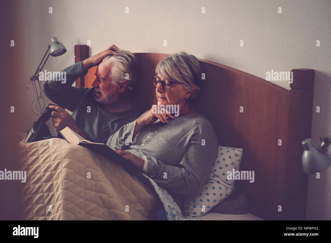 caucasian senior adult couple stay and rest on the bed. the wman read a book the man play with goggled headset technology. beautiful model and light f Stock Photo