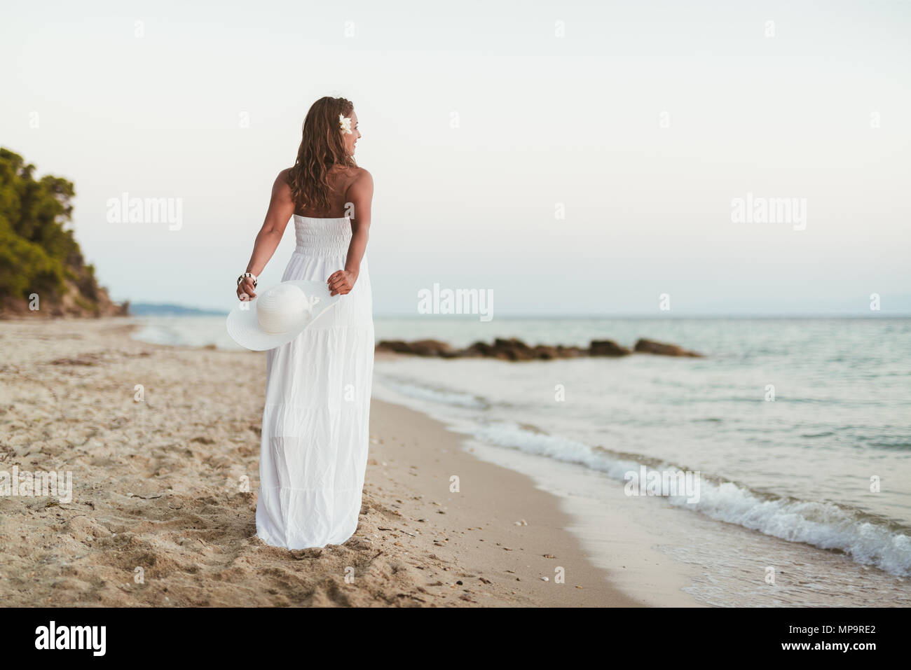 Rear view of a beautiful young smiling woman in white dress walking on ...