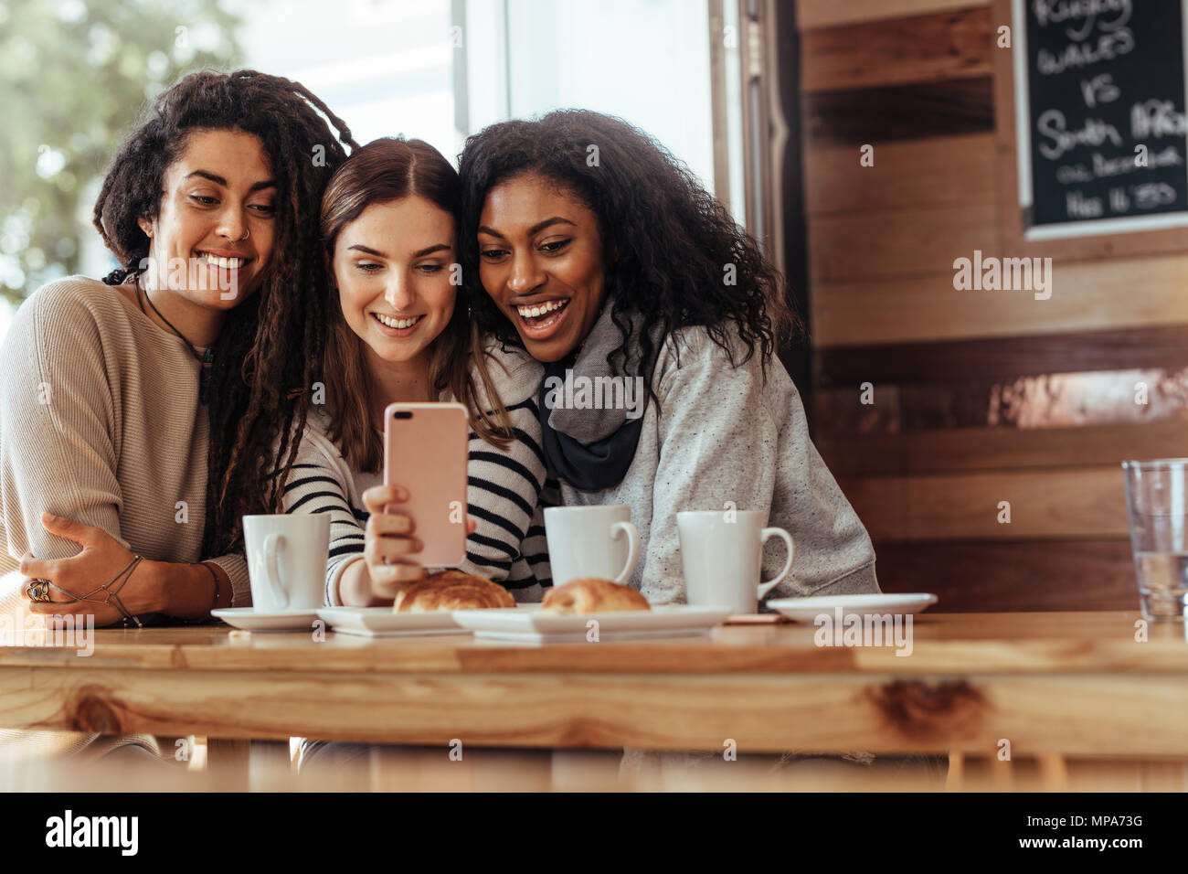 Three women sitting in a restaurant looking at mobile phone and smiling. Friends sitting at a cafe with coffee and snacks on the table looking at a mo Stock Photo