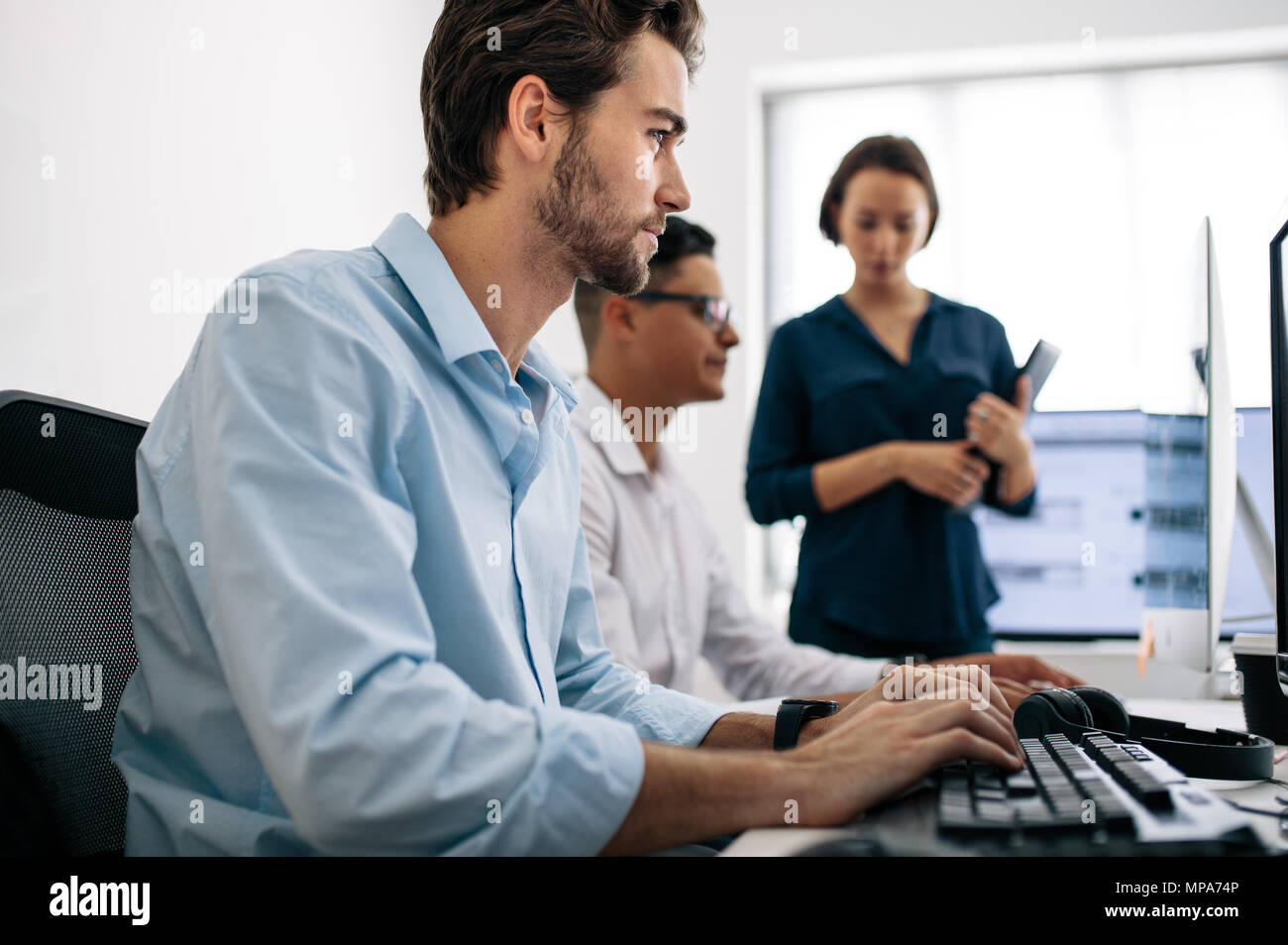 Software developers sitting at office working on computers. Two men developing applications on computer while a female colleague looks on. Stock Photo