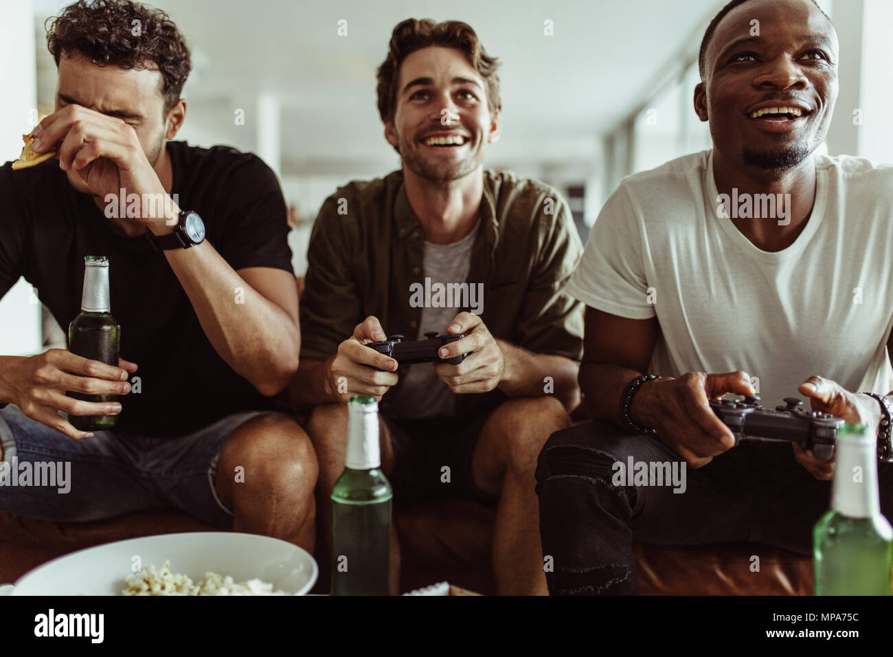 Friends having fun with drinks and snacks while playing video game at home. Two men playing video game holding joysticks while another man watches wit Stock Photo