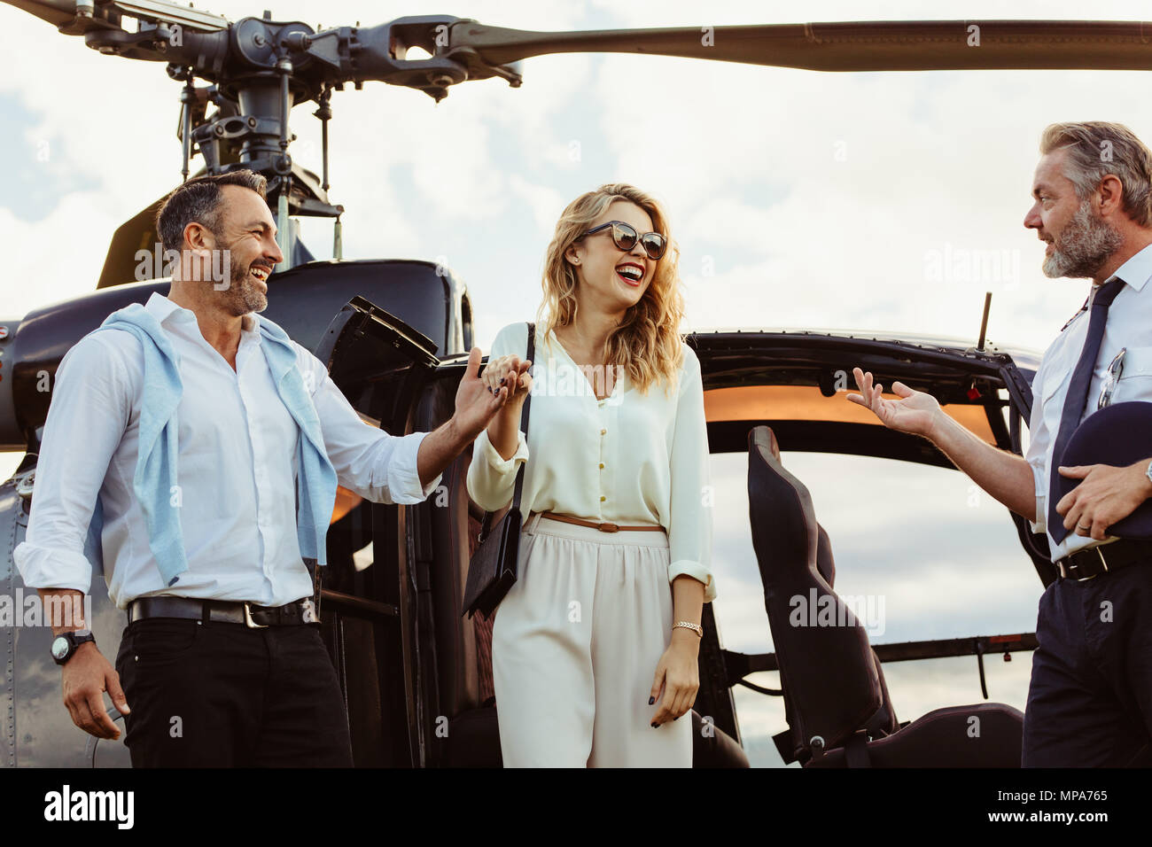 Smiling man and woman alighted from a private helicopter with pilot standing by. Couple getting off a private aircraft with mature pilot. Stock Photo