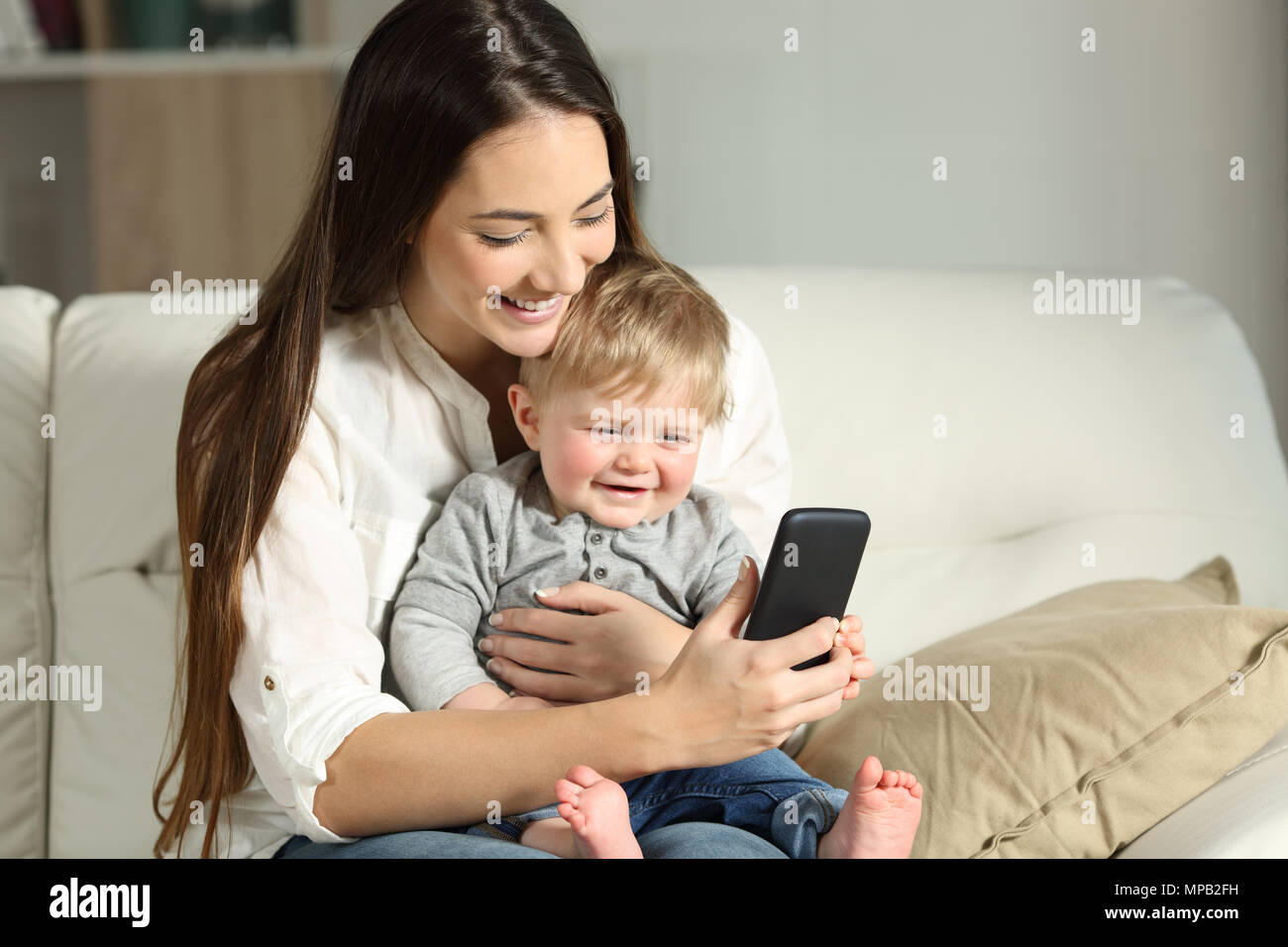 Mother and baby playing with a smartphone sitting on a couch in the living room at home Stock Photo