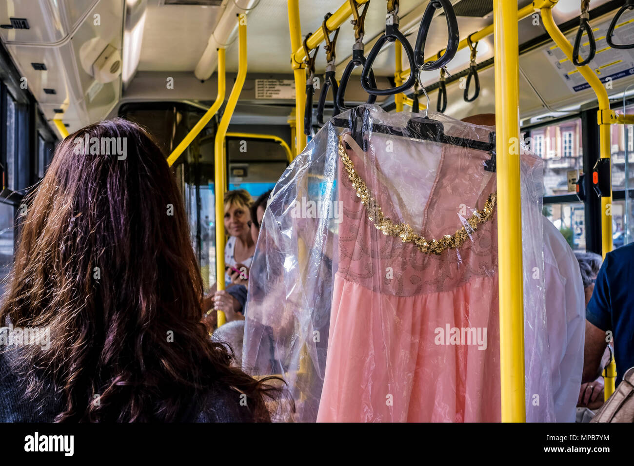 Woman fetched a pink dress with sequins (paillettes), wrapped in a dry cleaning laundry plastic bag, hung on handrail on a city urban public bus. Stock Photo