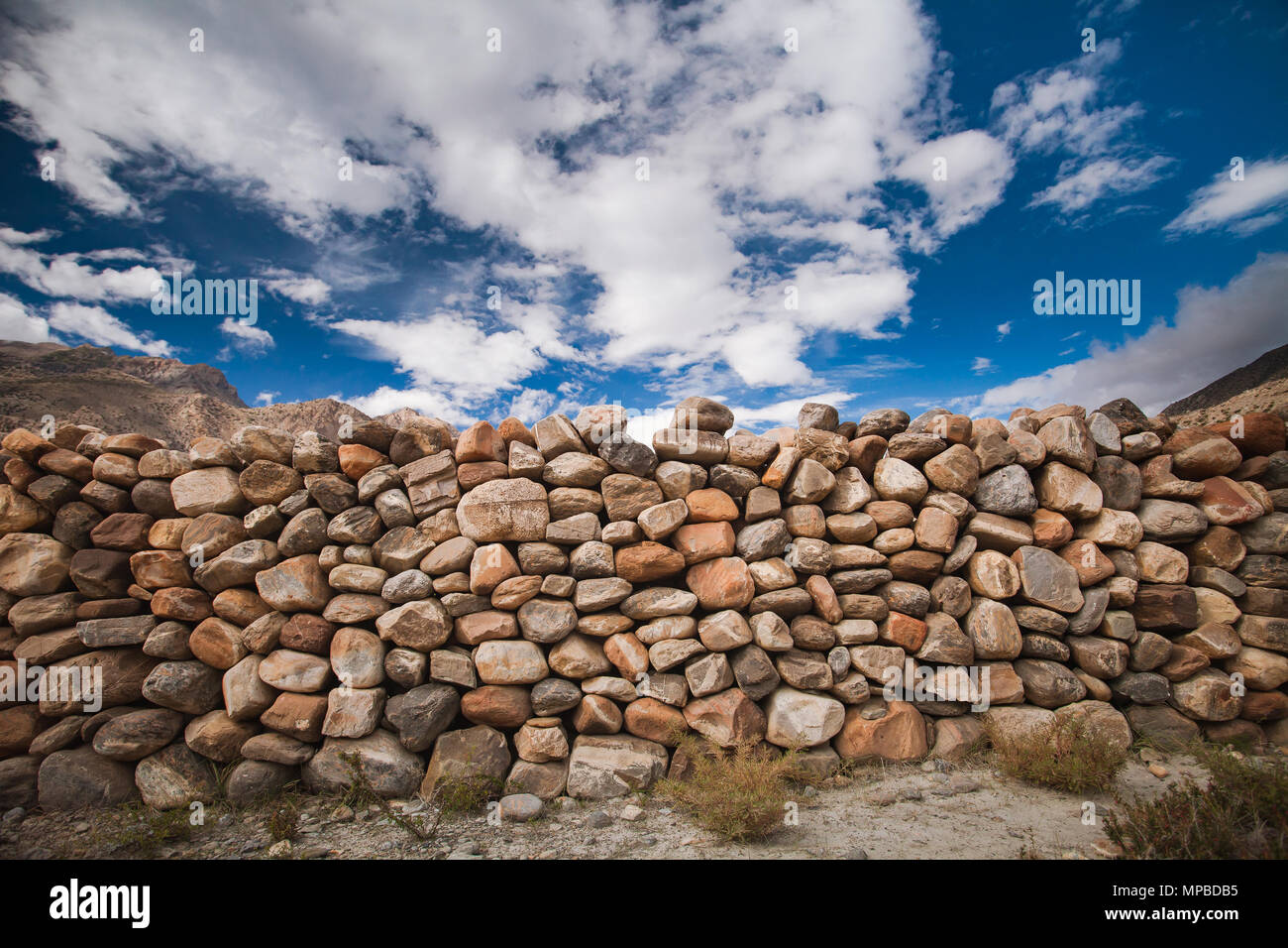 The stone wall constructed from the different sizes the boulders in grey, brown, tawny tints. Bright blue sky with white clouds. Ideal background for the illustrations and collages. Stock Photo
