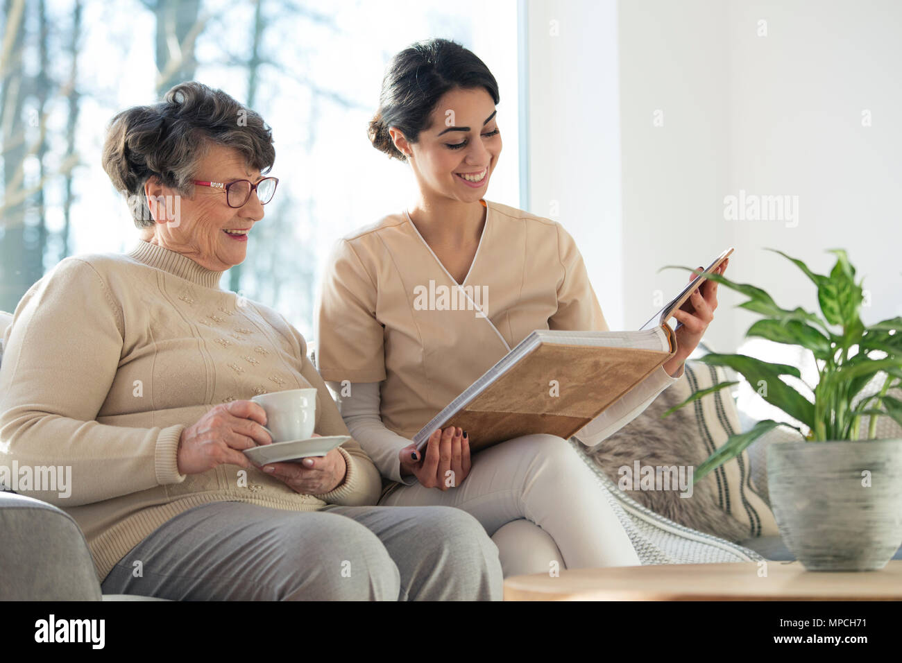 Smiling professional caretaker showing a family album to an elder woman in a living room at home Stock Photo