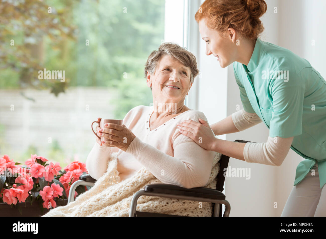 A professional caretaker in uniform helping a geriatric female patient on a wheelchair. Senior holding a cup and sitting by a large window in a rehabi Stock Photo