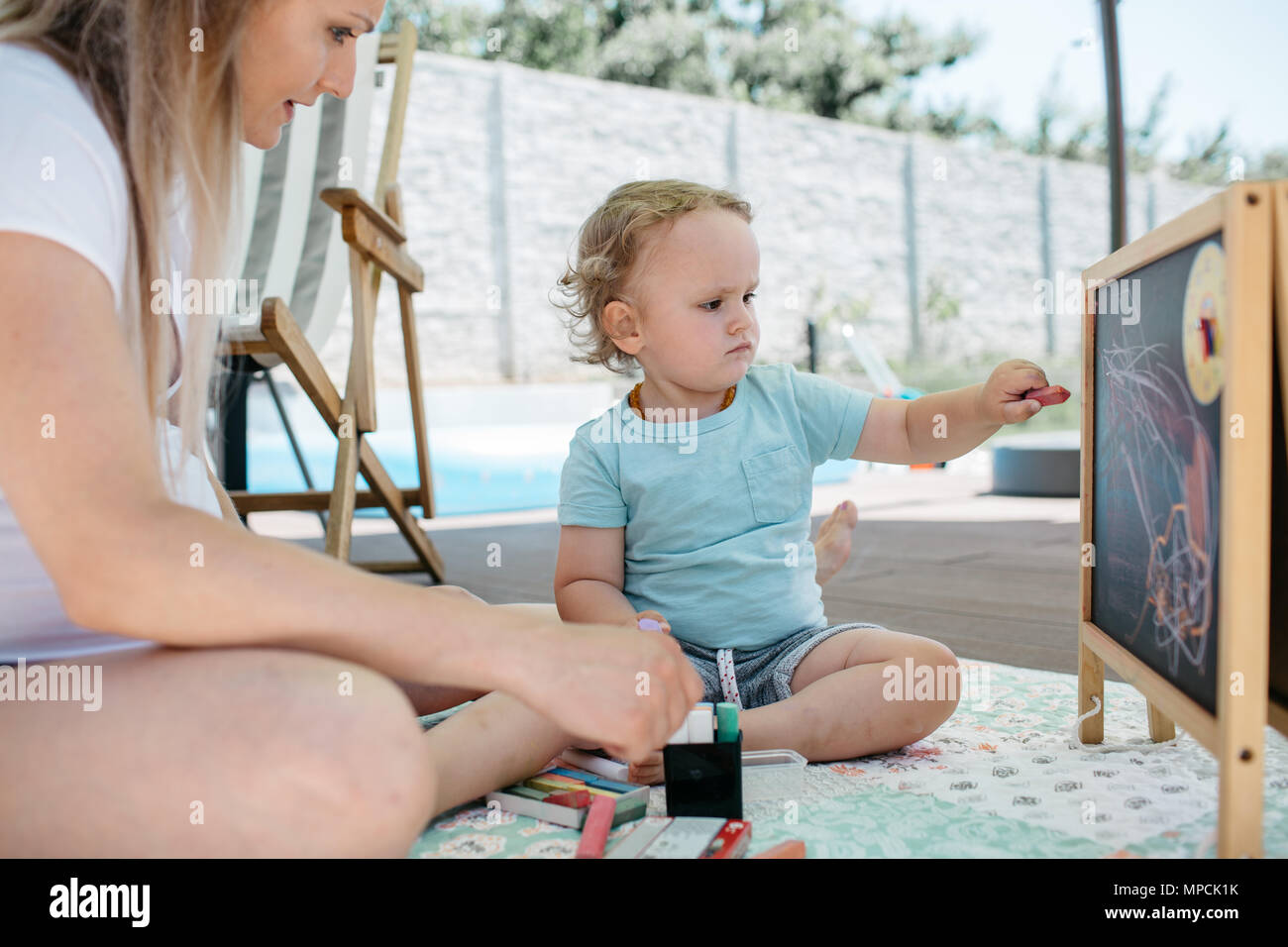 A little boy and his mother sitting on a patio and drawing on a chalkboard. A mother admiring first drawings of her son on a chalkboard. Stock Photo
