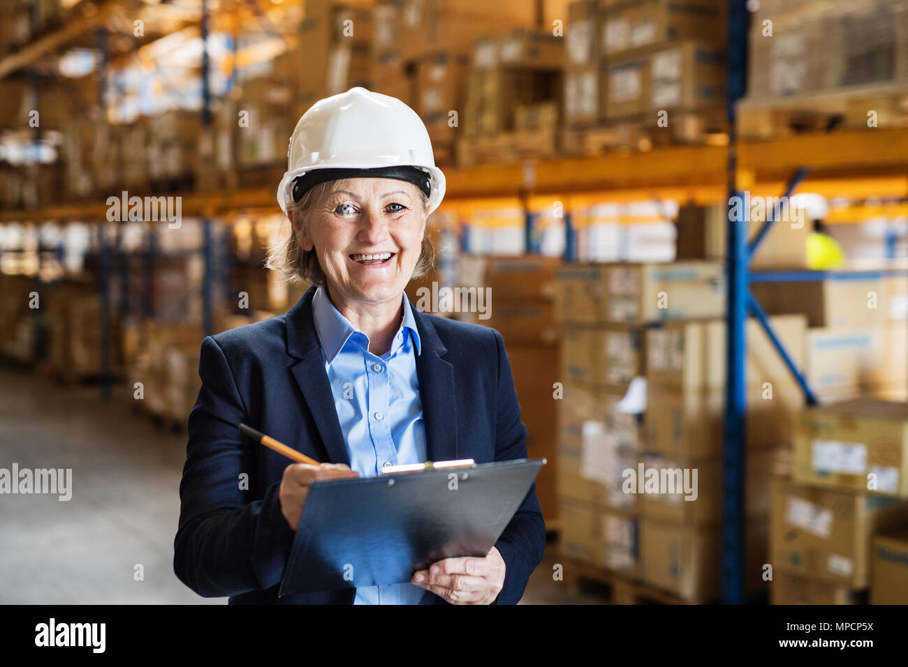 Senior woman warehouse manager or supervisor with white helmet and clipboard. Stock Photo