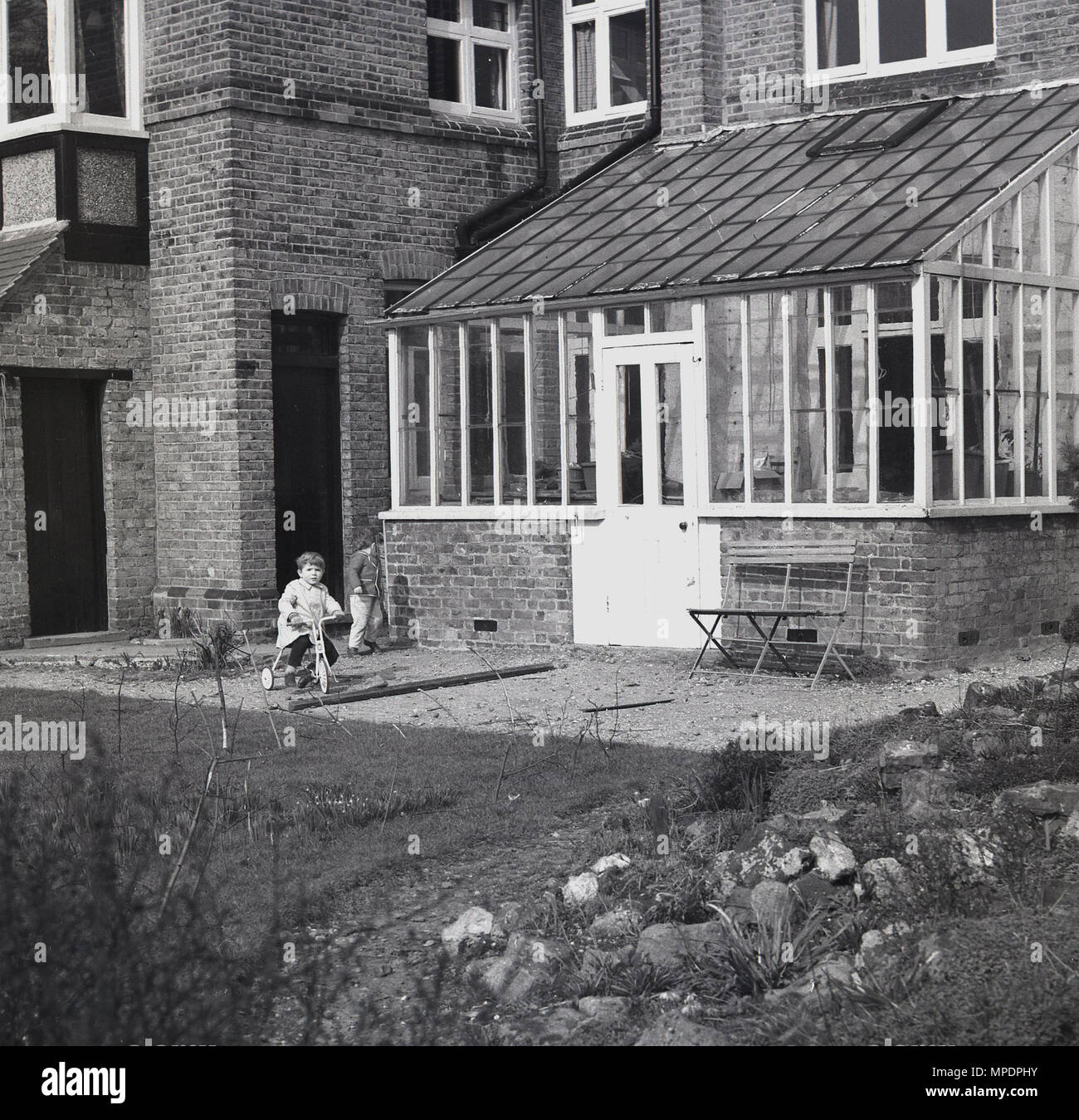 1961, historical, small boy rides a tricylce on an area of gravel in